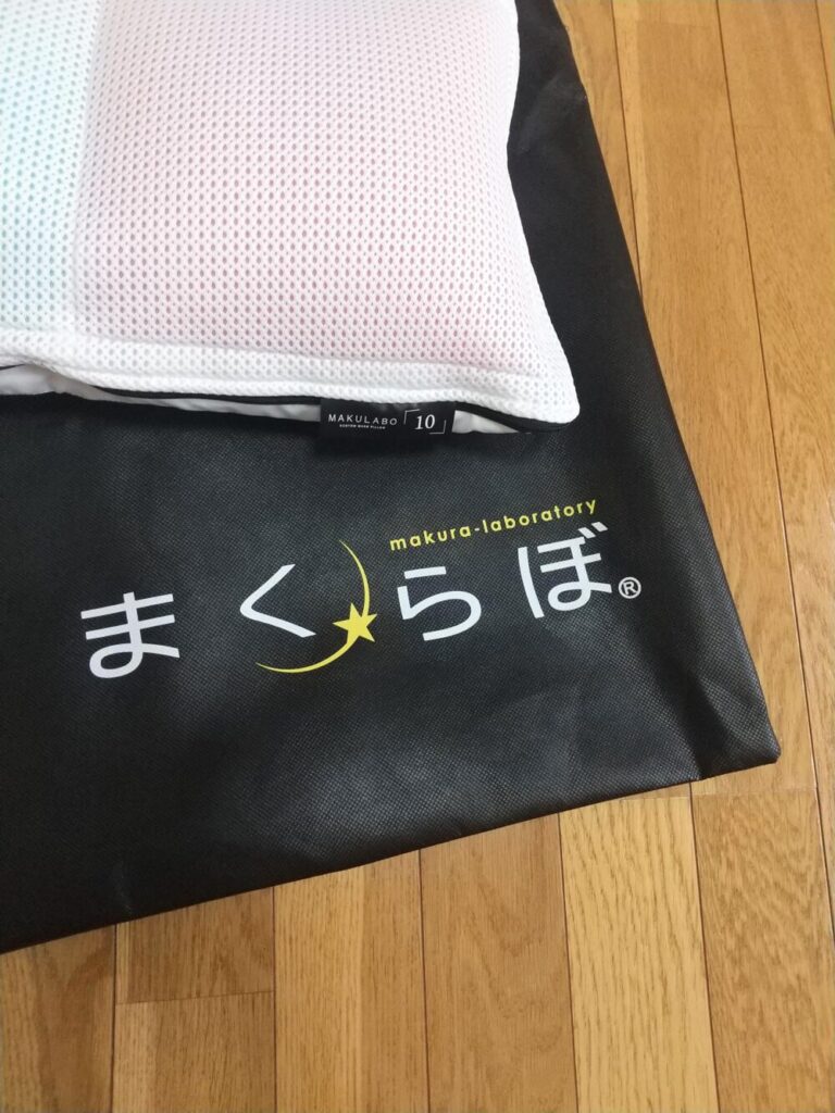 まくらぼのオーダーメイド枕「レギュラー」と、まくらぼのロゴが入った手提げ袋を床に並べた写真